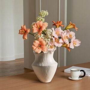 Modern off-white organic ceramic vase filled with peach and pink lilies resting on a wooden dining table beside a coffee cup