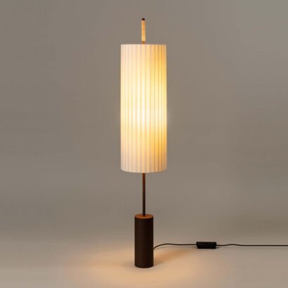A modern minimalist floor lamp with a illuminated tall cylindrical pleated white fabric shade, a slender dark metal pole, and a solid dark brown cylindrical wood base on a plain grey background.