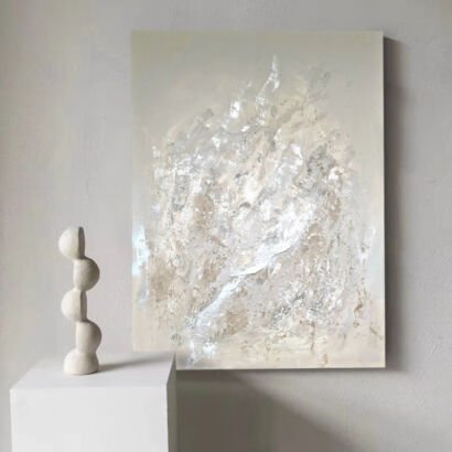 A vertical heavily textured abstract canvas painting featuring shimmering silver foil highlights layered over a soft cream and beige plaster background, displayed beside a white pedestal with a modern sculpture.
