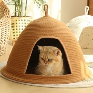 A short-haired golden tabby cat lounging inside a camel-colored woven cotton rope dome cat cave bed with a wide arched opening and top loop, set on a light-colored rug with plants and another white cat bed in the background.
