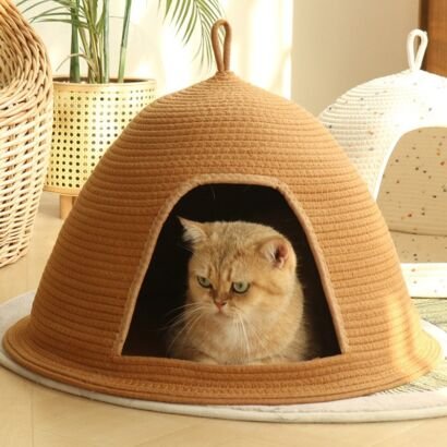 A short-haired golden tabby cat lounging inside a camel-colored woven cotton rope dome cat cave bed with a wide arched opening and top loop, set on a light-colored rug with plants and another white cat bed in the background.