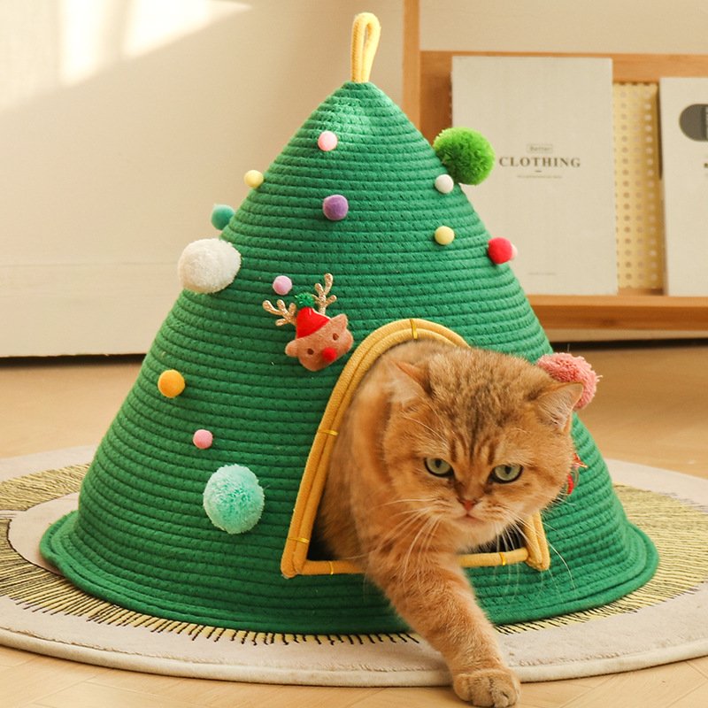 A ginger tabby cat stepping out from a green woven cotton rope Christmas tree-shaped cat cave bed, decorated with colorful pom-poms and a cute plush reindeer patch on a round rug.