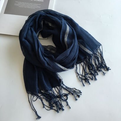 A navy blue lightweight linen scarf with subtle silver metallic lurex stripes and knotted fringe ends, artfully bundled on a white surface next to a magazine.