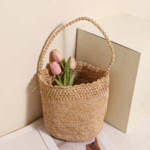A natural beige handwoven straw bucket tote bag filled with pink tulips, featuring a scalloped crochet rim and a single long braided top handle, set beside a white block against a neutral backdrop.