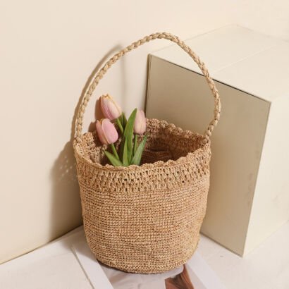 A natural beige handwoven straw bucket tote bag filled with pink tulips, featuring a scalloped crochet rim and a single long braided top handle, set beside a white block against a neutral backdrop.