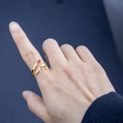 A close-up shot of a person's hand pointing upward while wearing an open-wrap gold ring on the index finger, featuring a personalized cursive name on one end and a round red birthstone on the other, set against a dark background.