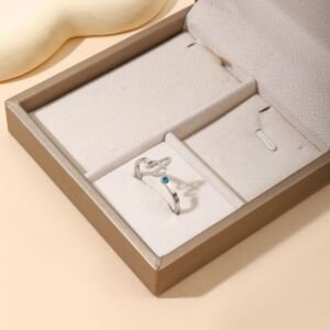 A delicate silver open wrap ring featuring a personalized cursive initial letter on one end and a bright blue round birthstone gem on the other, displayed inside a beige suede jewelry box insert.