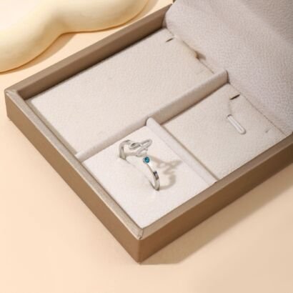 A delicate silver open wrap ring featuring a personalized cursive initial letter on one end and a bright blue round birthstone gem on the other, displayed inside a beige suede jewelry box insert.