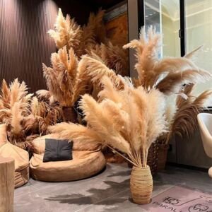 A dramatic, voluminous floor display of natural beige and taupe fluffy pampas grass plumes arranged in a ribbed woven rattan vase, set in a cozy modern lounge room with leather beanbag chairs and dark slatted wood accent walls.