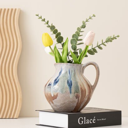 Handcrafted ceramic pitcher vase with blue and earthy brown drip glaze holding tulips and eucalyptus branches on a hardcover book
