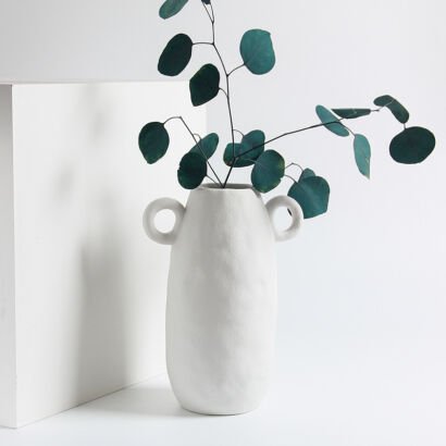 Matte white ceramic vase with round side loop handles holding fresh green eucalyptus branches on a white table