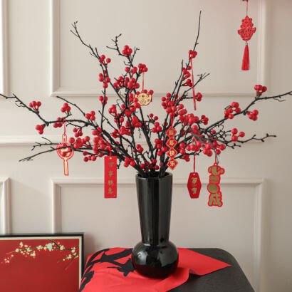 A black ceramic vase with a rounded base and slender neck filled with dark branches laden with vibrant red berries and hanging red festive ornaments, displayed on a table with a red cloth and a framed artwork against a neutral wall.