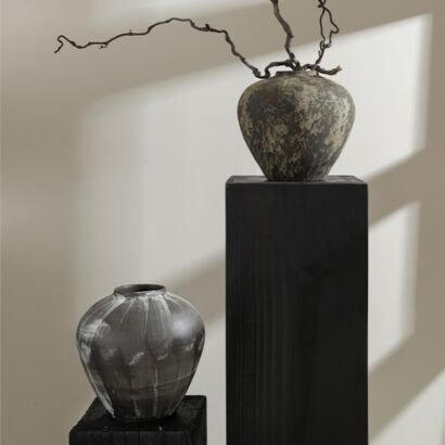 Two dark distressed ceramic vases with rustic textures displayed on black wood blocks, featuring a rounded dark vessel with mottled green-brown shading holding a winding dry branch on a tall pedestal block, alongside a smaller dark grey vase with subtle vertical brushstroke patterns.