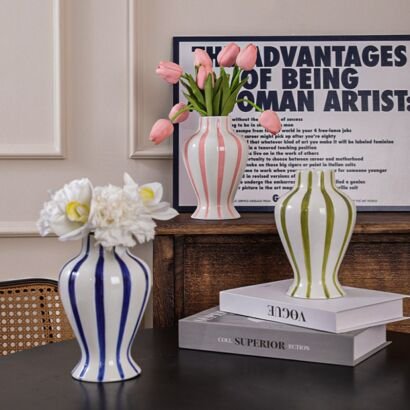 Three ceramic vases with vertical stripes in blue, pink, and green displayed together on tables and books, featuring rounded bodies, narrow necks, and flared rims, with pink tulips and white flowers arranged inside them.