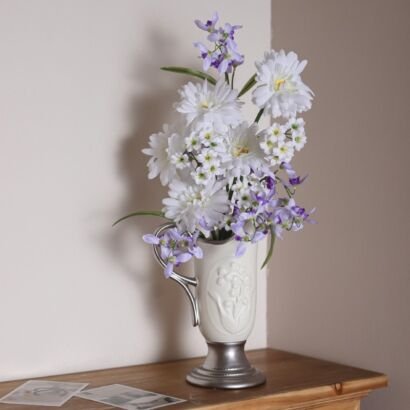 An elegant white ceramic pitcher-style vase with a silver-toned base, handle, and rim resting on a wooden tabletop against a corner wall, filled with a lush arrangement of white daisies and purple wildflowers, with small photographs lying on the surface beside it.