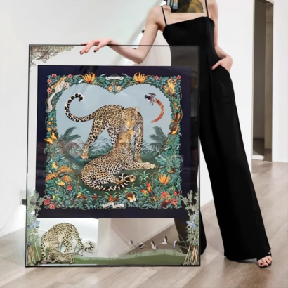 A large square transparent acrylic floating frame displaying a fine silk scarf with two leopards framed by tropical botanical flora, held next to a person in a black jumpsuit.