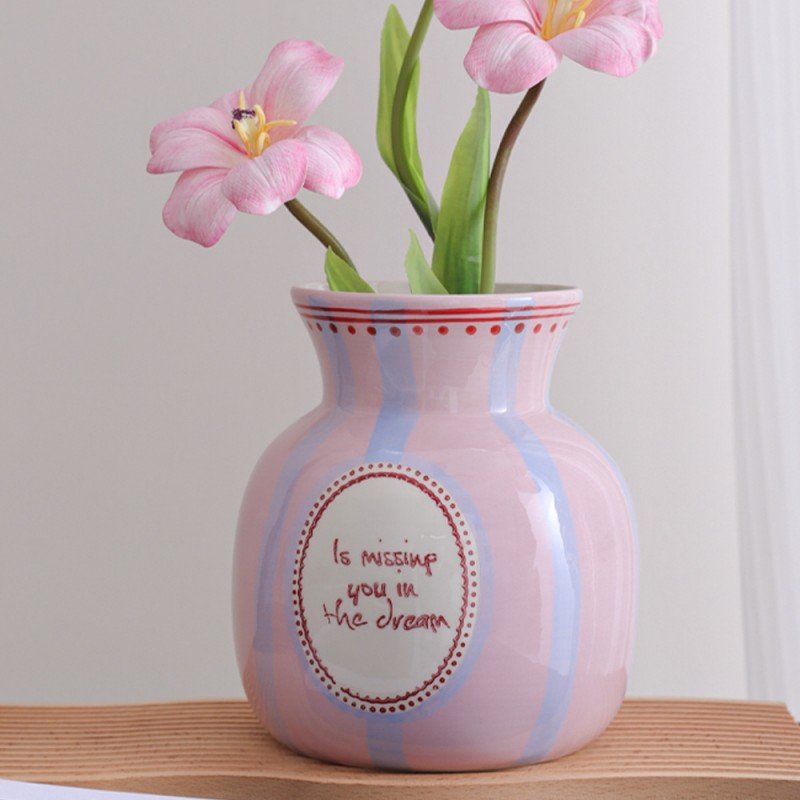 A round ceramic vase with pink and blue vertical stripes sitting on a wooden surface, featuring a white circular medallion on the front with the handwritten-style text "Is missing you in the dream", holding pink artificial flowers.