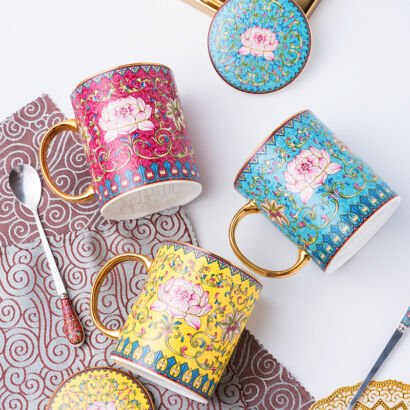 Three colorful ceramic coffee mugs with matching lids and spoons arranged on a white surface and patterned fabric, featuring intricate traditional Chinese cloisonné enamel lotus patterns in vibrant shades of pink, turquoise blue, and yellow with gold handles and rims.