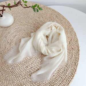 A soft cream white lightweight scarf with fine frayed raw fringe edges, artfully draped in a swirl on a woven round straw placemat next to a small white vase.