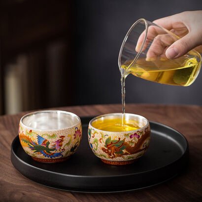 A hand pouring amber tea from a clear glass pitcher into one of two rounded ceramic teacups resting on a round black tray, showcasing detailed enamel dragon and phoenix motifs on the exterior and polished silver-lined interiors.