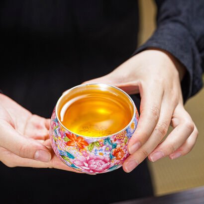 A person holding a handcrafted round ceramic teacup filled with clear golden tea, featuring a vibrant multi-colored enamel floral exterior with pink peonies and a silver-lined interior.