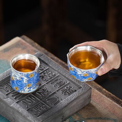 A person holding an octagonal handcrafted teacup filled with tea next to a second cylindrical teacup on a dark stone tile, both featuring a shiny silver-lined interior and vibrant blue wave enamel with golden blossom accents on the exterior.