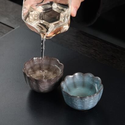 Clear tea pouring from a glass pitcher into one of two small handcrafted lotus flower-shaped ceramic teacups with scalloped rims and icy crackle glazes in slate blue and brownish grey on a dark stone tray.