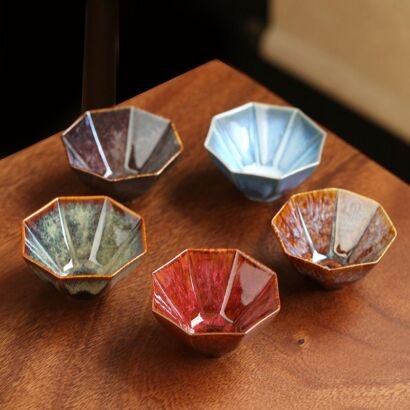 Five handcrafted hexagonal ceramic teacups with sharp geometric facets arranged on a warm wooden table, showcasing rich kiln-transformed reactive glazes in crimson red, amber brown, jade green, sky blue, and metallic bronze.