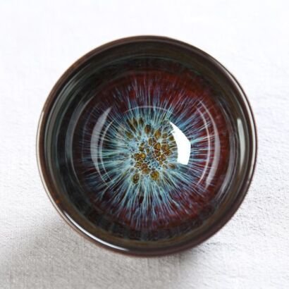 A top-down view of a small round ceramic Jian Zhan teacup revealing an intricate, starry kiln-transformed interior with iridescent blue rays radiating outward from a golden oil spot center.