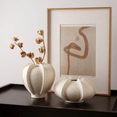 Two off-white textured ceramic vases with distinct vertical ribbed fins sitting on a dark table next to a framed minimalist abstract art piece