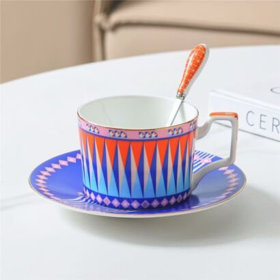 Vibrant porcelain teacup and saucer set featuring colorful orange, blue, and pink geometric triangular patterns resting on a white table with a matching ceramic spoon
