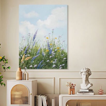A vertical canvas painting depicting a blooming wildflower meadow with purple lavender, yellow accents, and green grass under a bright blue cloudy sky displayed above a side table.