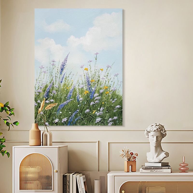 A vertical canvas painting depicting a blooming wildflower meadow with purple lavender, yellow accents, and green grass under a bright blue cloudy sky displayed above a side table.