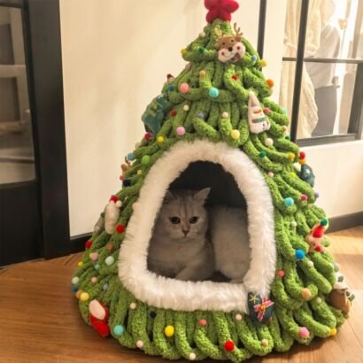 A fluffy grey cat resting comfortably inside a green hand-knitted Christmas tree-shaped pet bed decorated with colorful pompoms and festive ornaments