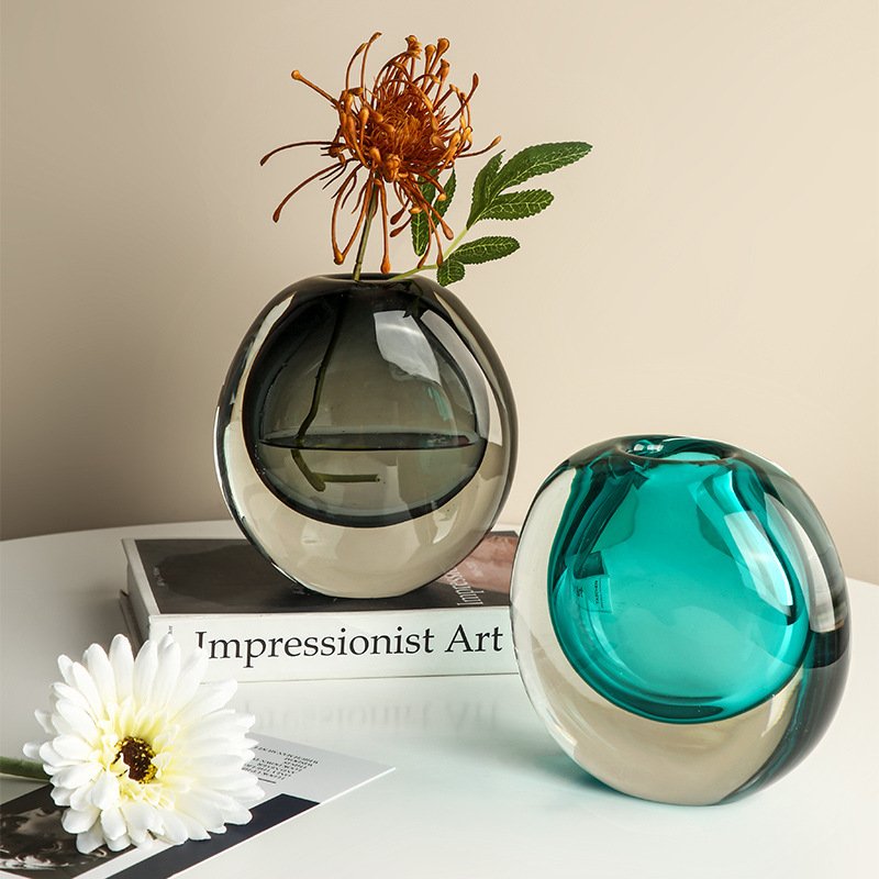 Two heavy round double-layer glass vases, one smoked grey with an orange spider lily flower and one vivid teal blue, sitting on a table next to an Impressionist Art book