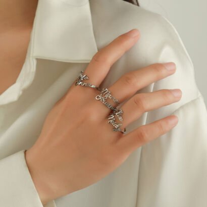 A close-up shot of a hand wearing a custom silver spiral wrap ring engraved with multiple personalized cursive names extending across the middle finger, set against a soft off-white collared shirt.
