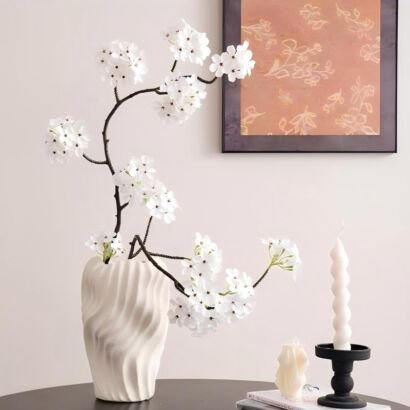A matte white ceramic vase with a dynamic vertical swirl texture filled with winding dark branches bearing delicate white cherry blossoms, displayed on a table next to a framed floral print, books, and spiral candles on holders.