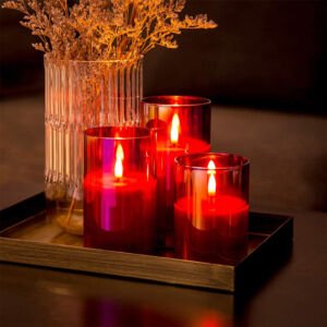 A cozy tabletop arrangement featuring a trio of red glass hurricane cylinder candles with warm flickering LED flames set on a metallic decorative tray next to a clear ribbed vase of dried flowers.