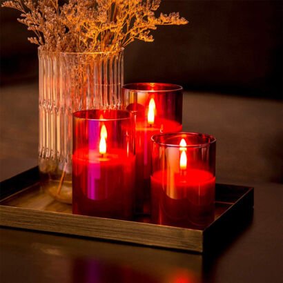 A cozy tabletop arrangement featuring a trio of red glass hurricane cylinder candles with warm flickering LED flames set on a metallic decorative tray next to a clear ribbed vase of dried flowers.