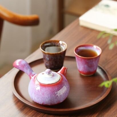 A handcrafted ceramic side-handle teapot with a vibrant magenta-purple kiln-transformed glaze and white lid, resting on a round wooden tray next to two small matching glazed teacups filled with tea.