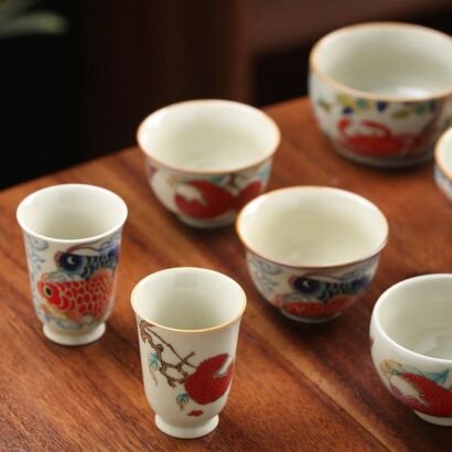 A selection of handcrafted ceramic teacups arranged on a warm wooden table, featuring handpainted traditional Chinese motifs like red koi fish, crabs, and lychee branches against a smooth ivory porcelain glaze.