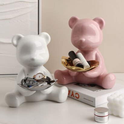 Two glossy ceramic bear statues in white and pink sitting on a table, holding metallic trays filled with keys, watches, and lipsticks