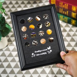 A small black framed shadow box displaying sixteen circular cutouts revealing colorful real butterfly wing patterns set against a black background with white cursive text.