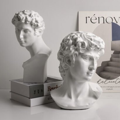 Two white resin Michelangelo David head bust statues sitting on a stack of decorative display books next to abstract art wall prints