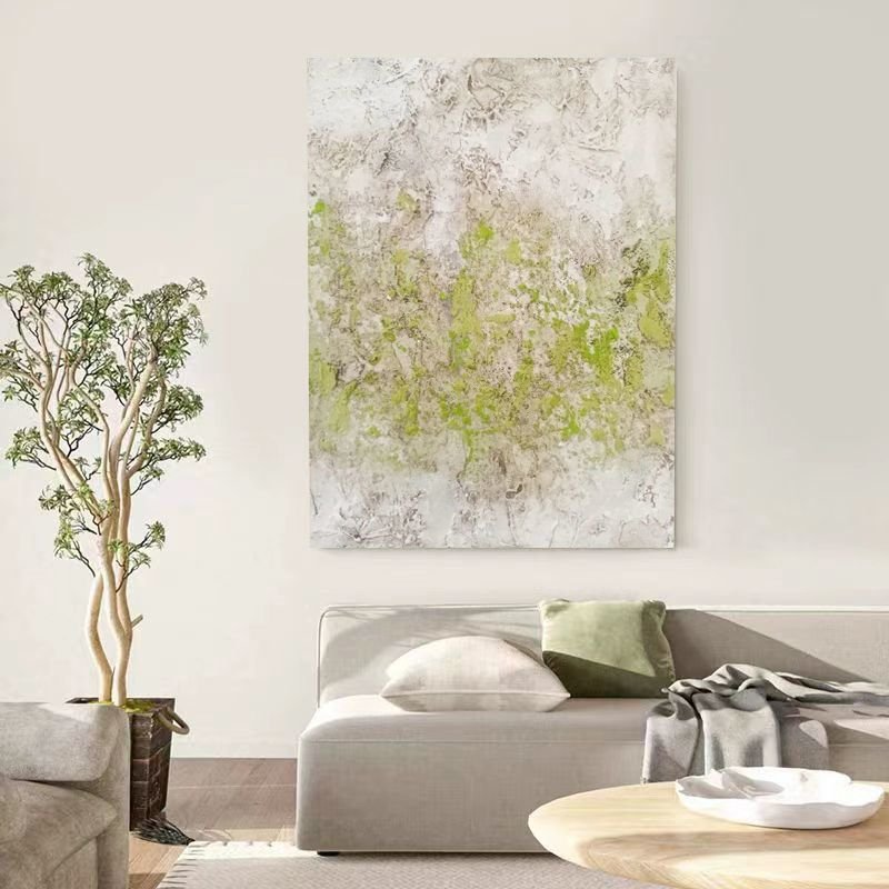 A vertical textured abstract canvas painting with subtle lime and moss green accents layered over a plaster-like beige background hanging above a modern couch in a neutral living room