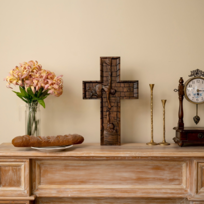 A dark wood carved wall cross displayed upright on a light wooden mantelpiece between a vase of pink flowers, a loaf of bread, two gold candle holders, and a vintage clock, featuring a high-relief stone-brick texture with figures carved into the cross.