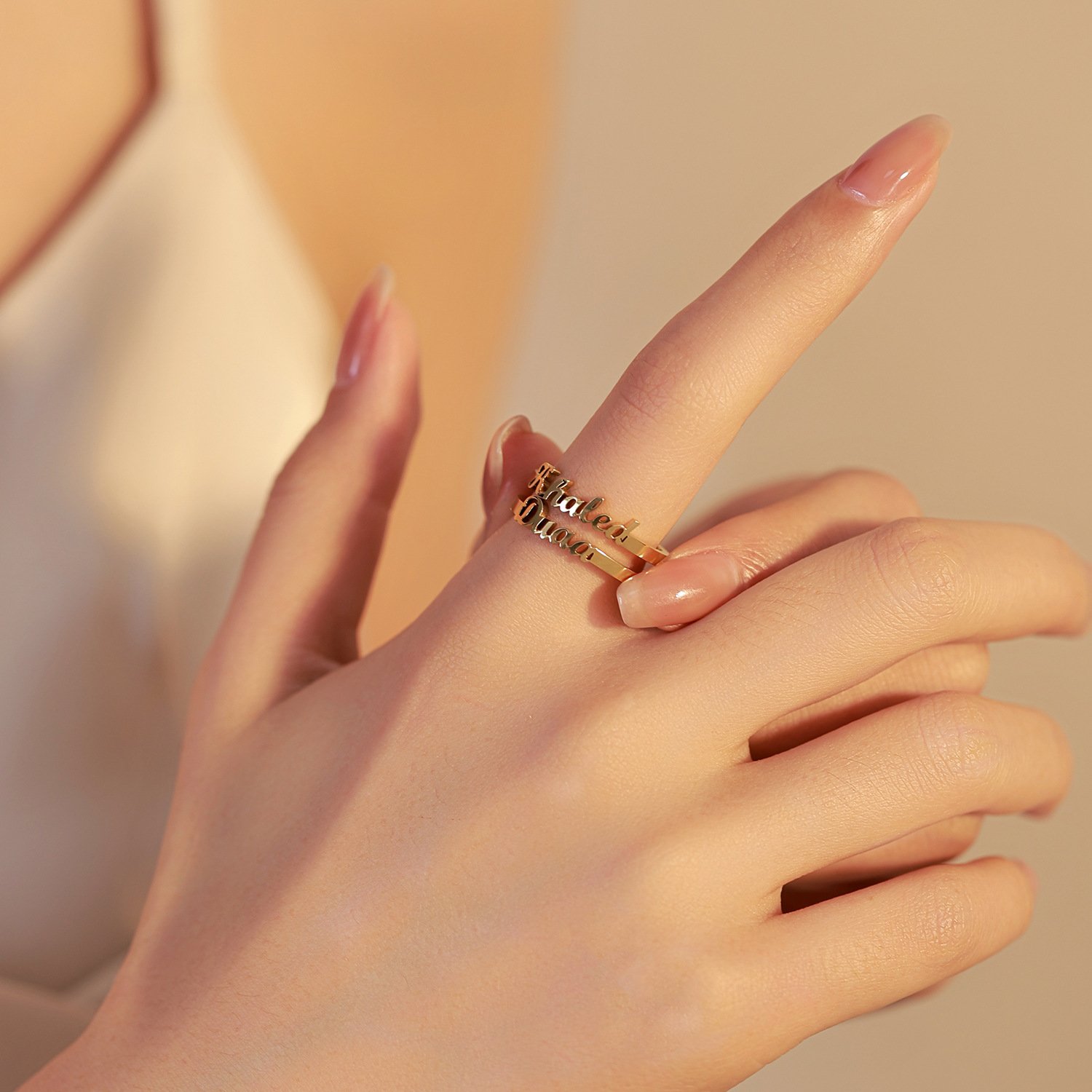 A close-up shot of a hand with nude manicured nails wearing a personalized double-layer gold ring with script cursive names on the index finger against a warm, soft-lit background.