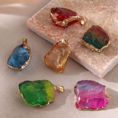 A collection of six colorful raw crystal gemstone nugget pendants in red, iridescent rainbow, ocean blue, honey yellow, emerald green, and magenta pink, each framed with textured gold electroplated edges and fitted with a gold bail on a stone and sandy surface.