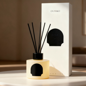 A frosted pale yellow glass reed diffuser bottle with a black collar and arched label, holding five black diffuser sticks, sitting next to its minimalist white tall rectangular packaging box in a warmly lit room.
