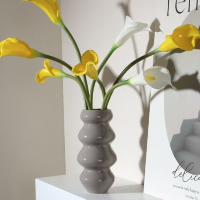 A glossy grey ceramic vase with a unique stacked bubble or gourd-like segmented silhouette, displayed on a white shelf and filled with an arrangement of bright yellow and white calla lilies.